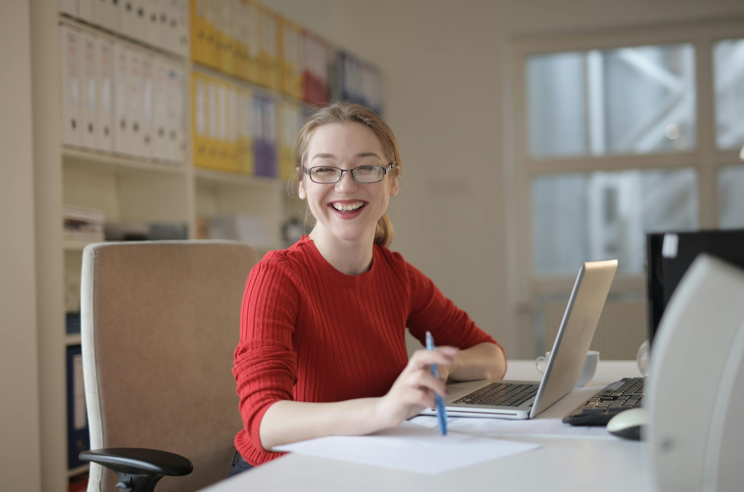 smiling woman wearing a red sweater, sitting at a desk with a laptop